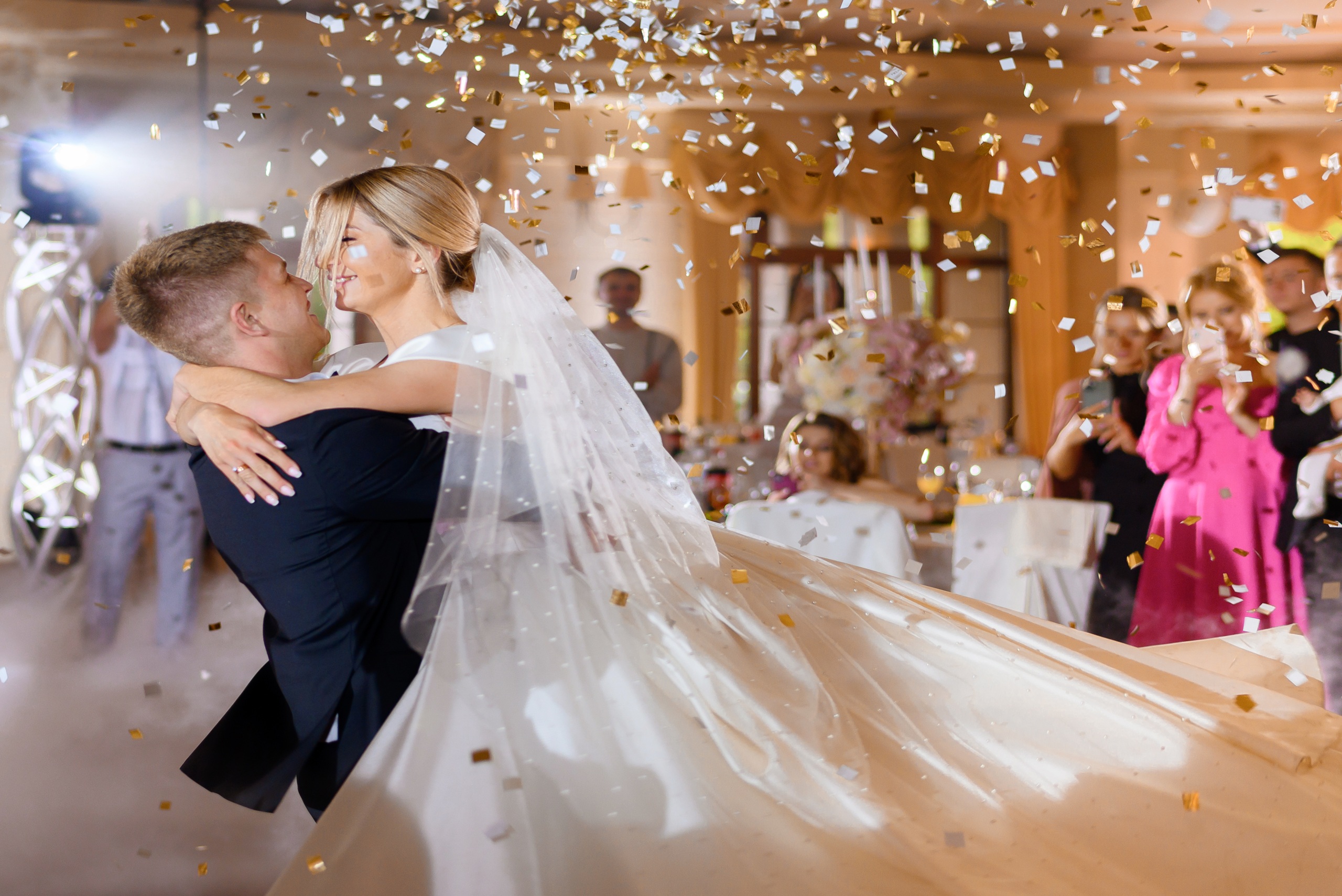 Side view of cheerful groom rising pretty bride in long wedding dress and veil, loving man waving girl and dancing in restaurant while falling confetti