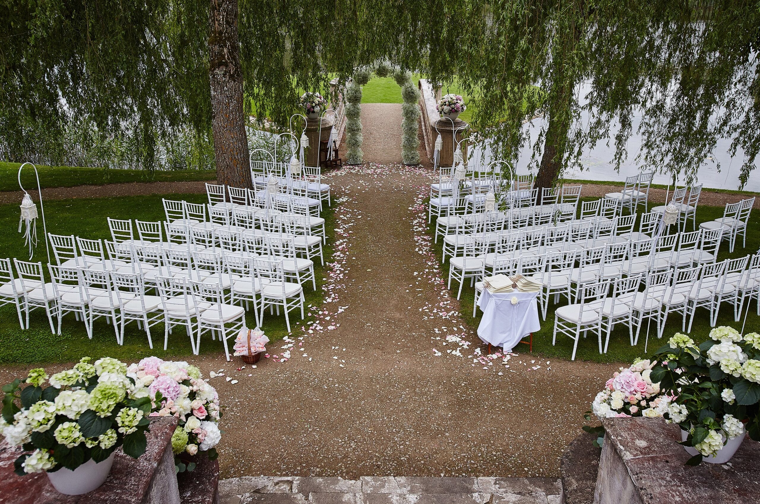Place for wedding ceremony with wedding arch decorated with flowers and white chairs on each side of archway outdoors. Preparation for wedding ceremony outdoors near lake.